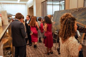 Guests at a formal event mingle and admire artwork on display. Several people, dressed in elegant attire, engage in conversation while others observe paintings set on easels. The setting is a well-lit indoor venue with a sophisticated atmosphere.