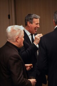 A group of well-dressed men in suits enjoying appetizers at a formal event, with one man laughing while taking a bite. The atmosphere appears lively and social.