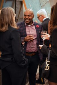 A man in a checkered blazer with a red poppy pin is smiling and holding a wine glass while engaged in conversation with a group of people at a formal event. Other attendees in elegant attire are mingling in the background.