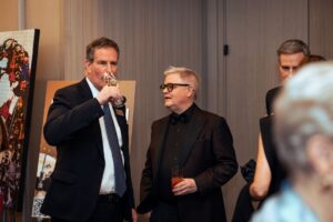 Two men in formal attire engaged in conversation at a networking event. One man in a dark suit sips from a wine glass, while the other in a black tuxedo with a bow tie holds a cocktail. A colorful painting is visible in the background.