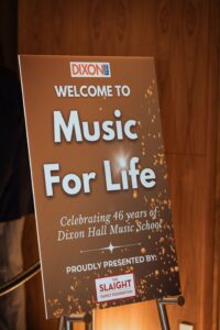 A promotional sign for the "Music For Life" event, celebrating 46 years of Dixon Hall Music School. The sign features a warm golden design with sparkling details and includes the event's title, a welcome message, and sponsorship information.