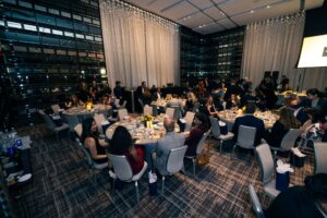 A banquet hall filled with elegantly set tables and guests dressed in formal attire. The room features floor-to-ceiling windows and decorative hanging lights, creating a sophisticated atmosphere. Attendees are engaged in conversation while enjoying their meals.