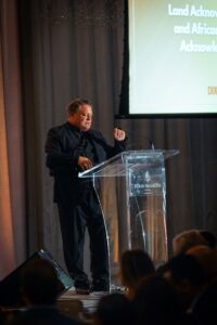 A man dressed in black is speaking at a clear podium with a microphone at the Four Seasons Hotel Toronto. A screen displaying event details is visible in the background, and audience members are seated in the foreground.