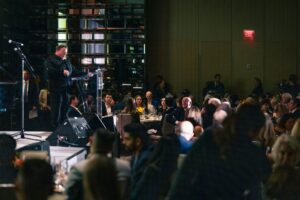 A speaker at a podium addressing a seated audience at a formal dinner event. The dimly lit room has elegant table settings, floral centerpieces, and guests attentively listening.