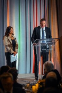 A man in a suit speaks at a podium with the Four Seasons Hotel Toronto logo, while a woman in a beige blazer stands beside him holding papers, smiling. The background features colorful drapery and an audience watching the presentation.