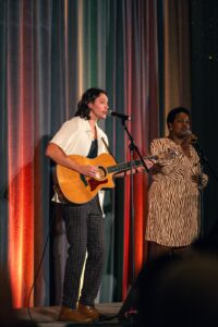 A male guitarist and a female singer perform on stage, both singing into microphones against a colorful draped backdrop. The guitarist wears a white shirt and plaid pants, while the singer wears a patterned dress.
