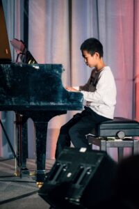 A young boy in a white long-sleeve shirt and black pants playing a grand piano on stage. He is focused, with his fingers on the keys, and a curtain backdrop is visible behind him.